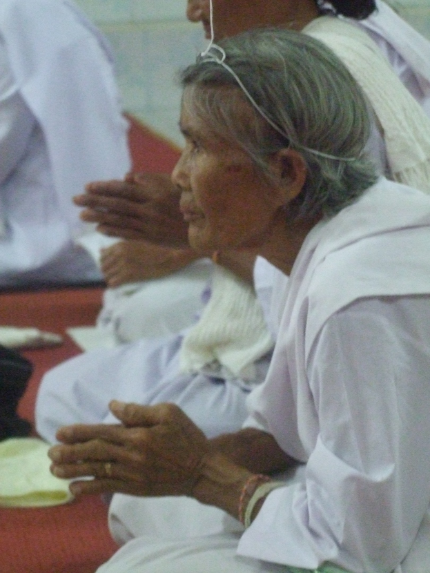 Alte Frau im Krankenhausbett liegend mit gefalteten Händen, geschlossene Augen und Kopf auf den Händen ruhend, umgeben von Menschen im Hintergrund.