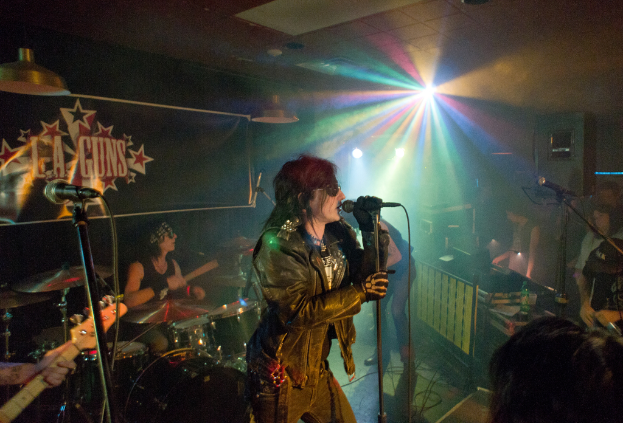Eine Frau in einer schwarzen Jacke singt auf der Bühne in ein Mikrofon, während ein Mann mit Sonnenbrille und Headband hinter ihr eine Gitarre spielt; im Hintergrund sind ein 'LA Guns'-Plakat und Diskoleuchten zu sehen.