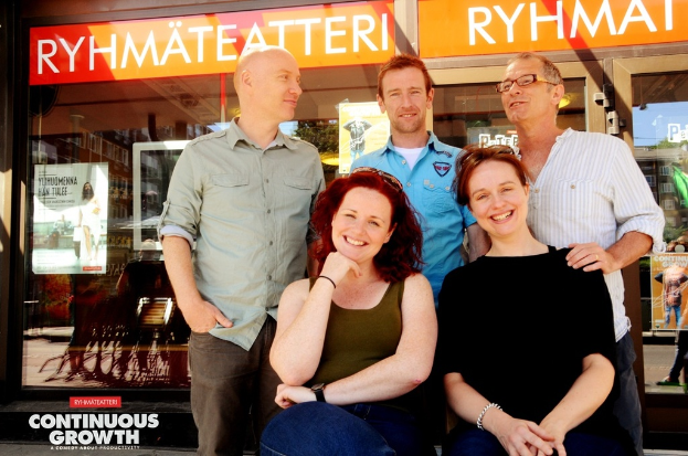 Drei Männer stehen und zwei Frauen sitzen vor ihnen, alle lachend, mit Geschäften im Hintergrund, die Brillengeschäfte anzeigen, und einem Schild mit der Aufschrift 'RHYME TEATTERI' über einem der Geschäfte.