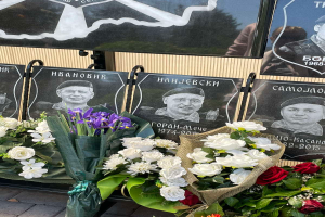 Ein Strauss bunter Blumen liegt auf einem Gehweg neben einem Schild, im Hintergrund sind mehrere Gedenktafeln zu sehen.