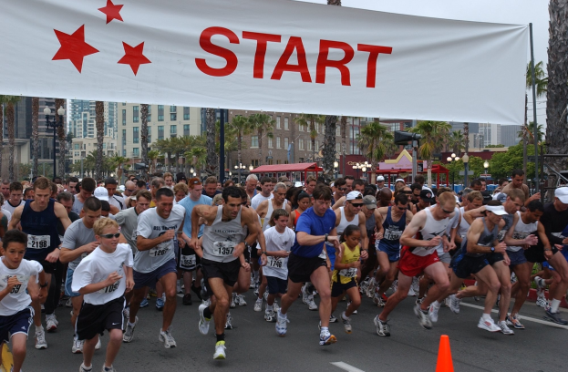 Gruppe von Menschen beim Marathon mit einem Verkehrskegel im Vordergrund und einem Banner mit Text im Hintergrund, umgeben von Bäumen, Laternenmasten, Gebäuden und einem klaren blauen Himmel.