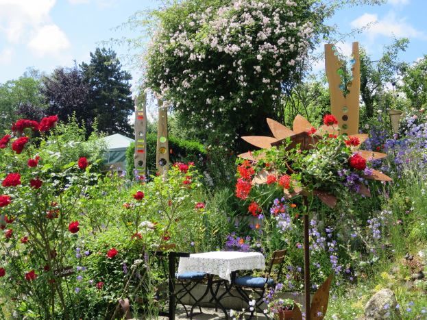 Ein Garten auf der Chelsea Flower Show mit bunten Blumen und Pflanzen, einem Tisch mit einer Decke und Stühlen in der Mitte und Bäumen im Hintergrund gegen einen bewölkten Himmel.