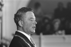 Ein Mann im Anzug und Krawatte steht an einem Podium mit einem Mikrofon und spricht zu einer unscharfen Menge, wobei er wie ein Politiker wirkt.