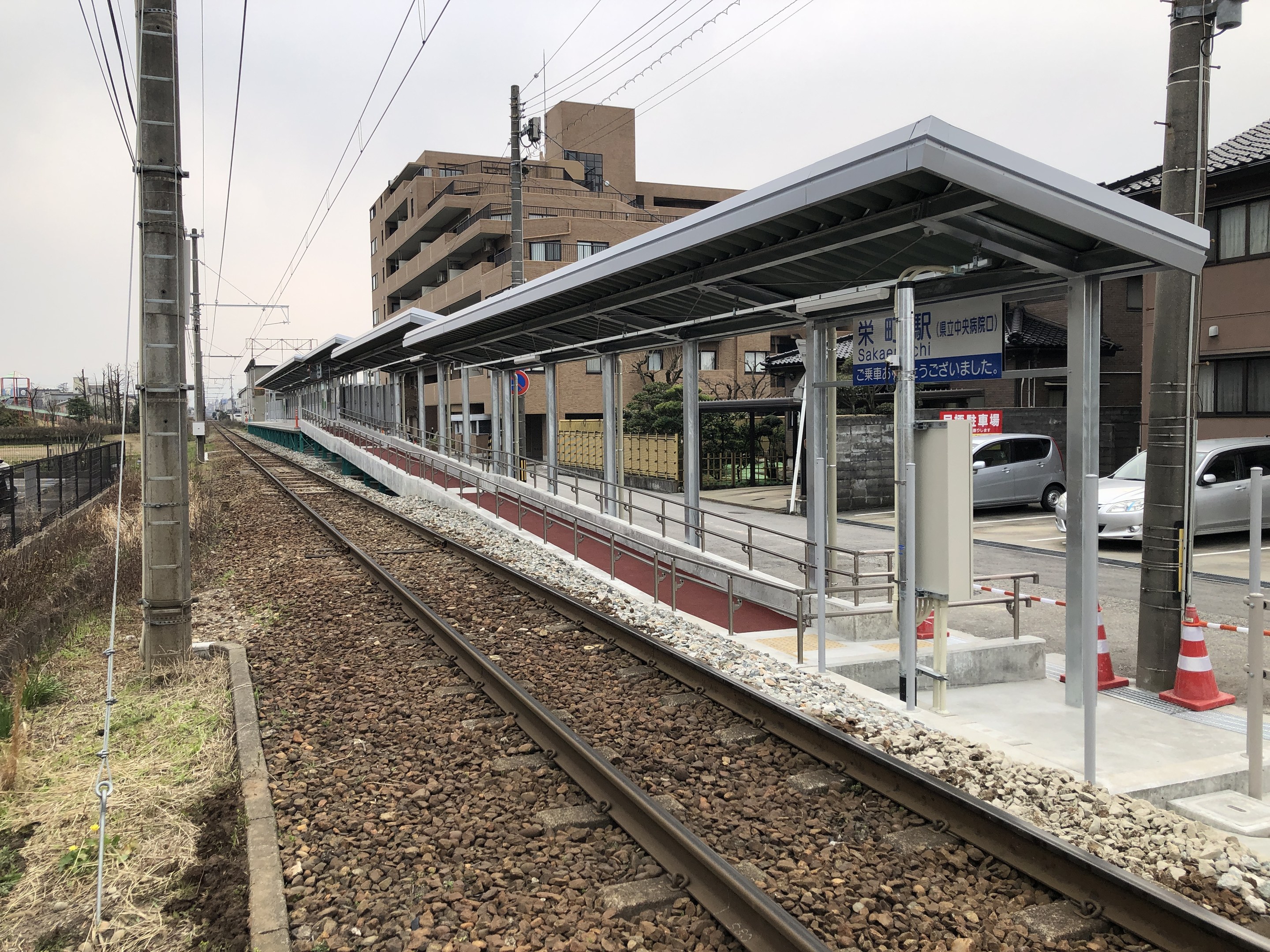 Ein Bahnhof mit einem Zug auf den Schienen, umgeben von Gebäuden, Strommasten, Kabeln, Fahrzeugen, Verkehrskegeln, Infotafeln, Zäunen, Bäumen und einem klaren blauen Himmel.