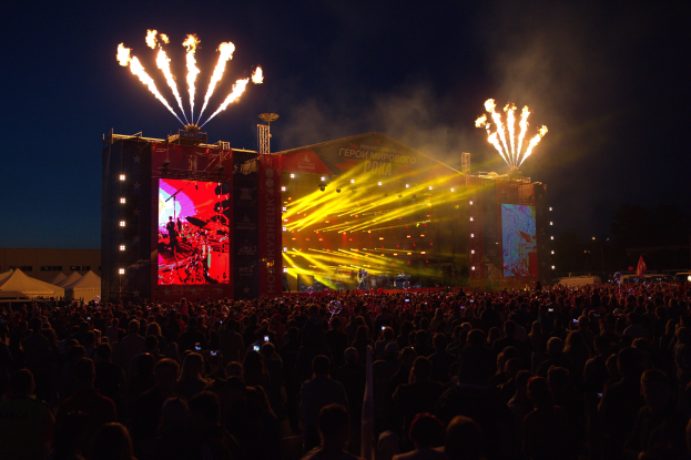 Eine große Menschenmenge auf einem Musikfestival schaut auf der beleuchteten Bühne mit Bannern, Zelten links und Feuerwerk am dunklen Himmel.