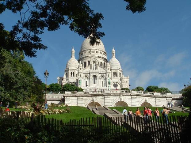 Ein weißes Gebäude umgeben von einem Zaun, Gras, Bäumen und Menschen mit Stangen und verschiedenen Pflanzen in der Nähe.