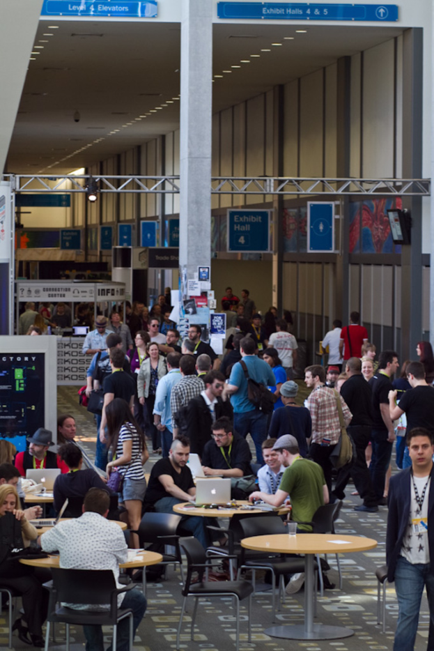 Ein belebter Flughafenempfang mit wartenden Menschen, einige stehen und andere arbeiten, Richtungsschilder an der rechten Wand und oben sowie von der Decke hängende Lampen.