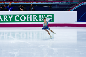 Eine Frau in einem blauen Kleid schlittert über eine Eisbahn, während eine Menge mit Kameras zusieht, mit einem Schild im Hintergrund, auf dem 'Tessa Virtue und Scott Moir' steht.