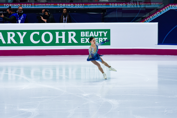 Eine Frau in einem blauen Kleid schlittert über eine Eisbahn, während eine Menge mit Kameras zusieht, mit einem Schild im Hintergrund, auf dem 'Tessa Virtue und Scott Moir' steht.