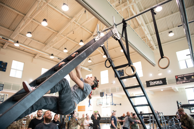 Ein Mann, der Klimmzüge an einer Leiter in einem Fitnessstudio macht, umgeben von Zuschauern mit Kameras, mit Fitnessgeräten und Bannern im Hintergrund.