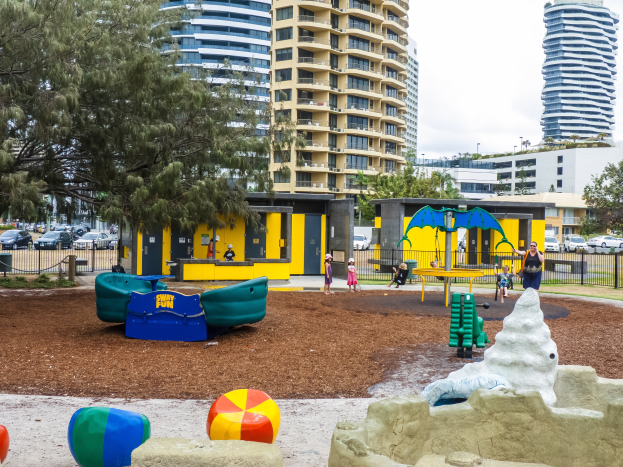 Ein Stadtspielplatz mit Kindern auf der Ausrüstung, umgeben von Bäumen und Geländern, mit hohen Gebäuden und Fahrzeugen auf der Straße im Hintergrund, unter einem sichtbaren Himmel.
