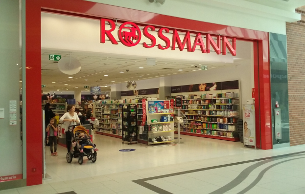 Ein Rossmann-Laden in einem Einkaufszentrum mit Menschen auf dem Boden, einer mit einem Kinderwagen, und Regalen voller verschiedener Artikel unter Deckenleuchten und Textschildern.