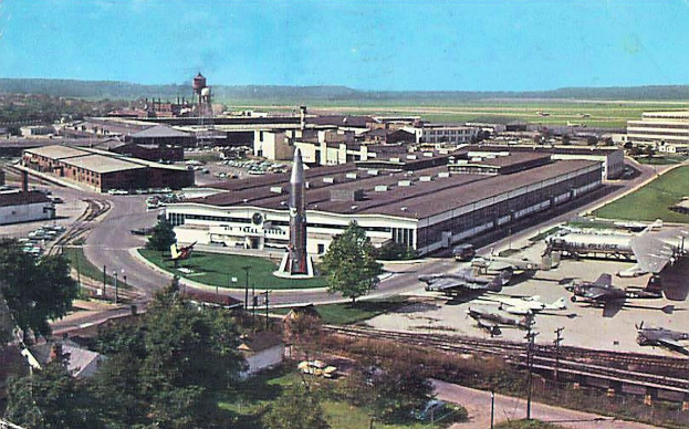 Altes Schwarz-Weiß-Foto eines Flughafens mit parkenden Flugzeugen, Gebäuden, Bäumen, Straßen, Fahrzeugen, Strommasten, Gras, Bahnschienen, Türmen, fernen Hügeln und einem klaren blauen Himmel, mit Text unten.