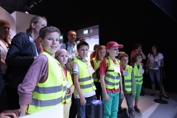 Eine Gruppe von Kindern mit Sicherheitswesten und Namensschildern, wobei ein Junge auf der linken Seite lächelt, auf einem Boden mit einem Pfahl auf der rechten Seite und einigen Menschen im Hintergrund steht.