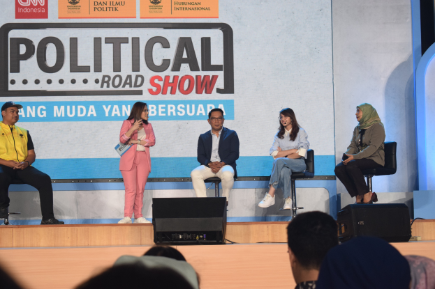 Eine Gruppe von Menschen sitzt auf einer Bühne mit einer Frau, die ein Mikrofon hält, in der Mitte, einem Publikum im Vordergrund und einem Banner mit der Aufschrift "Politischer Roadshow 2019" im Hintergrund.