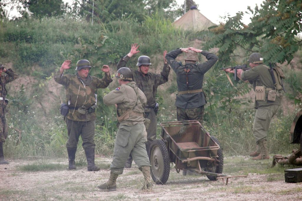 Mehrere Personen in Militäruniformen und Helmen stehen neben einem Wagen, umgeben von Pflanzen, Bäumen und Gras unter einem klaren Himmel.