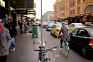 Ein Mädchen mit Helm fährt Fahrrad auf einer vielbefahrenen Straße mit vielen Fahrzeugen, Gebäuden auf der rechten Seite, Fußgängern auf der linken Seite, einem geparkten Fahrrad auf dem Gehweg und beleuchteten Geschäften.