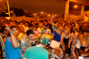 Menschen tanzen im Vordergrund bei einem Konzert, mit Lichtern, Pfählen, Bäumen und anderen Gegenständen im Hintergrund.