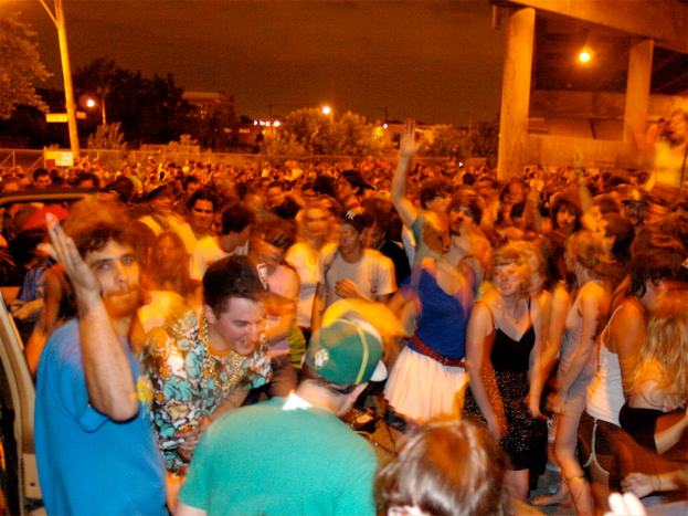 Menschen tanzen im Vordergrund bei einem Konzert, mit Lichtern, Pfählen, Bäumen und anderen Gegenständen im Hintergrund.