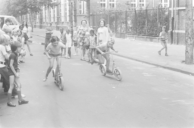 Schwarzes Bild von Kindern, die auf Rollern eine Straße entlangfahren, mit einem geparkten Auto auf der linken Seite, Gebäuden, Bäumen, Pfählen und einem Zaun im Hintergrund.
