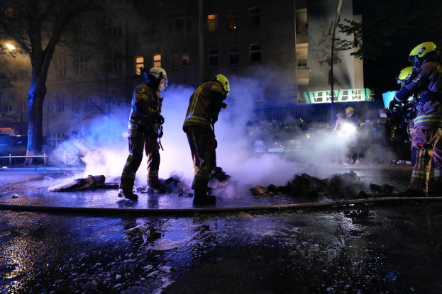 Feuerwehrleute in Helmen stehen nachts auf einer Straße mit Rauch aus einem Rohr aufsteigend, Gegenstände haltend, während Gebäude, Bäume und Fahrzeuge im Hintergrund zu sehen sind mit einer Tafel auf der rechten Seite.
