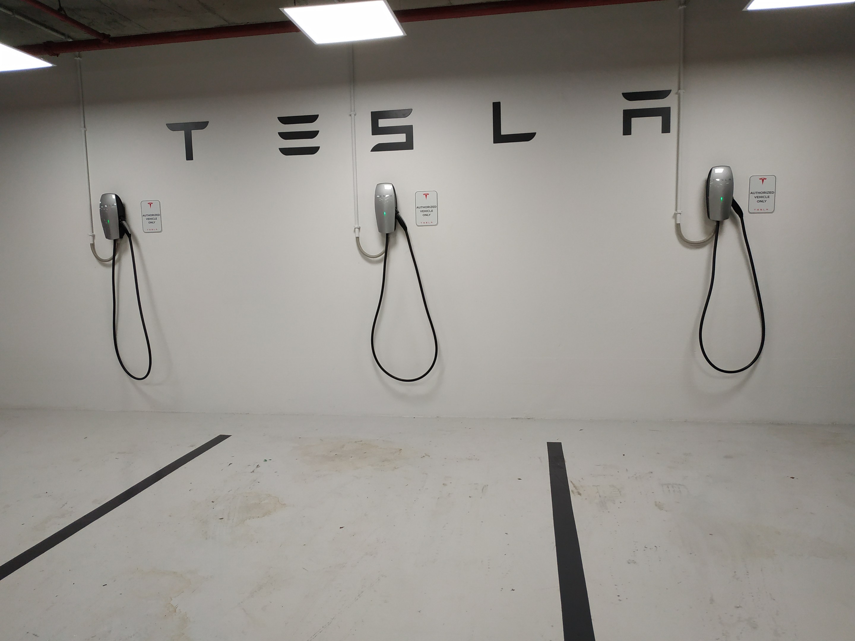 Eine Tesla-Ladestation in einer Tiefgarage mit angebrachten Kabeln und Deckenleuchten, die die darunterliegende Etage zeigen.