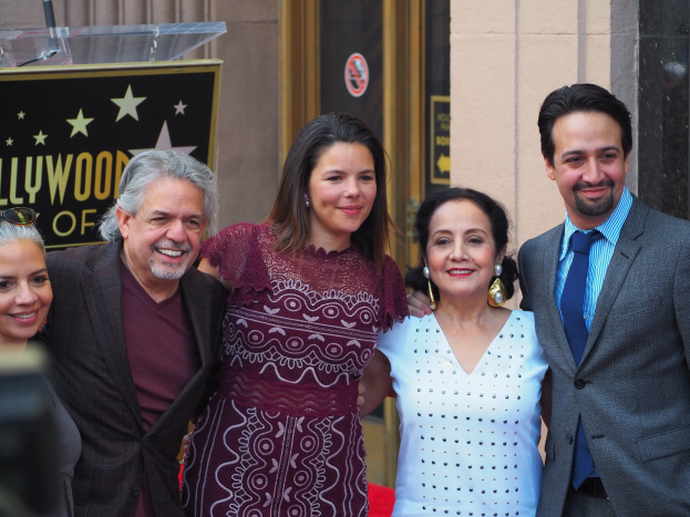 Gruppe von Menschen auf einem roten Teppich bei der Zeremonie des Hollywood Walk of Fame posend, mit einer Kamera auf der linken Seite und einer Tafel, einem Podium und einer Wand im Hintergrund.