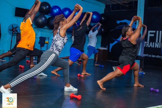 Gruppe von Menschen, die Yoga in einem Fitnessstudio mit Hanteln auf dem Boden machen, im Hintergrund ein Banner mit der Aufschrift 'Fitness-Training in Ghana'.