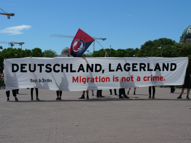 Eine Gruppe von Menschen hält ein Banner mit der Aufschrift "Deutschland, Lagerland Migration ist kein Verbrechen" und eine Flagge, mit Straßenlaternen, Bäumen, Kränen, einem Gebäude und einem bewölkten Himmel im Hintergrund.