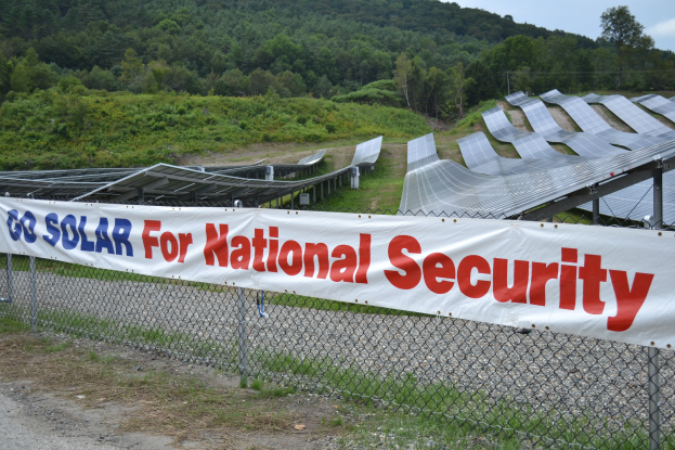 Schild mit der Aufschrift "Gehen Sie Solar für die nationale Sicherheit" vor einem Solarfeld mit Bäumen, Pflanzen und einem klaren blauen Himmel.