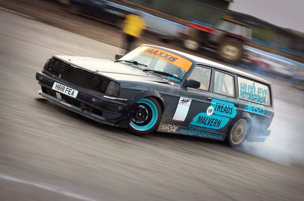 Ein Auto, das auf einer Rennstrecke fährt und Rauch hinter sich herzieht, umgeben von anderen Fahrzeugen und Zuschauern.