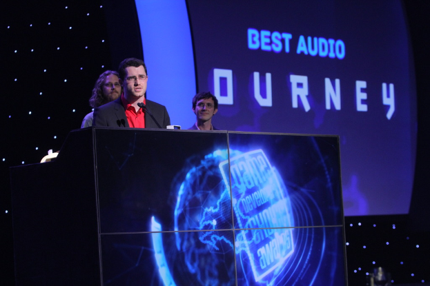 Drei Personen stehen auf einem Podium in einem Konferenzsaal mit einem Bildschirm und Lichtern im Hintergrund.