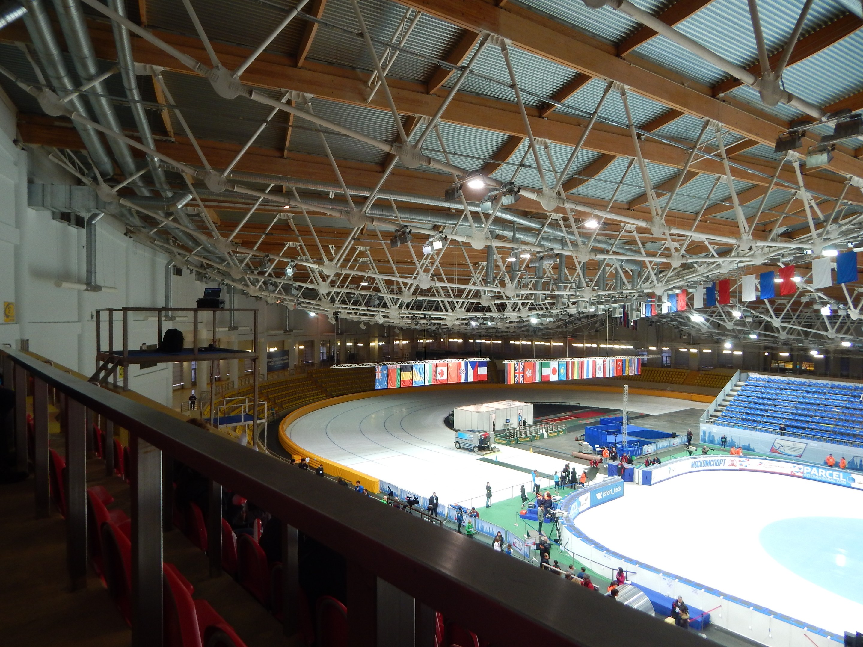 Große Indoor-Eisbahn mit zahlreichen Menschen, die Schlittschuhlaufen, umgeben von Sitzplätzen, Geländern, Brettern, Beleuchtungskörpern und strukturellen Elementen, mit einer Wand im Hintergrund.