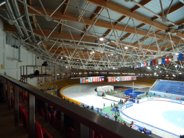 Große Indoor-Eisbahn mit zahlreichen Menschen, die Schlittschuhlaufen, umgeben von Sitzplätzen, Geländern, Brettern, Beleuchtungskörpern und strukturellen Elementen, mit einer Wand im Hintergrund.