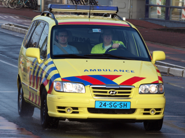 Ein gelber Krankenwagen mit zwei sichtbaren Insassen fährt an parkenden Fahrrädern auf einem Fußweg vorbei, mit einem Glasfrontgebäude im Hintergrund.