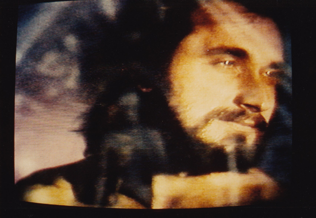 Ein Mann mit langem dunklem Bart und Haar, der ein weißes Hemd trägt, hat einen ernsten Gesichtsausdruck und schaut direkt in die Kamera auf einem Fernseher mit dunklem Hintergrund.