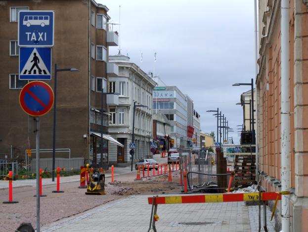 Stadtstraße mit Gebäuden, Straßeninfrastruktur, Fahrzeugen, einer Baustelle mit Verkehrszeichen, Bäumen und einem bewölkten Himmel.