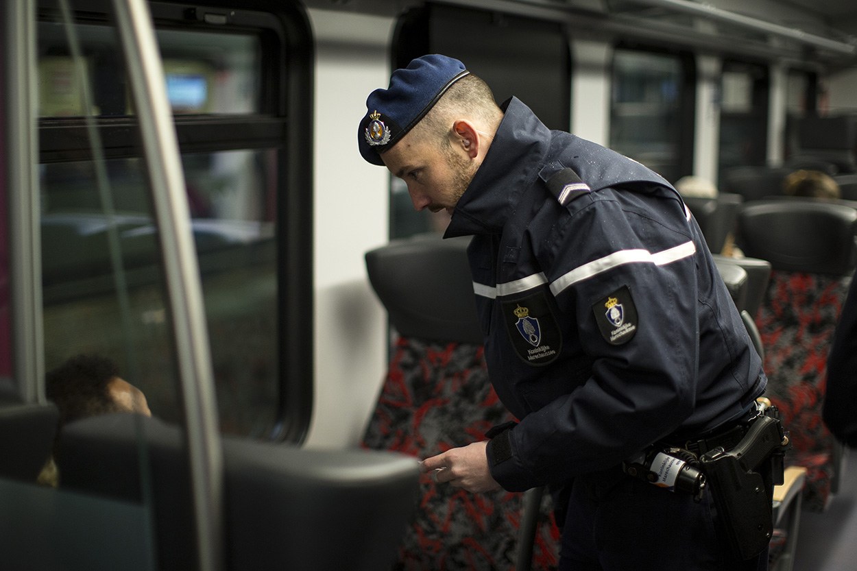Ein Polizist in Uniform und Mütze steht in einem Zug, schaut auf sein Handy, mit sitzenden Passagieren und einer weiteren stehenden Person im Bild.