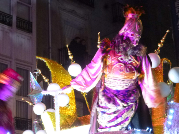Ein Mann in einem Kostüm steht auf einem dekorierten, beleuchteten Festwagen in einer Stadtumzug, mit einem Gebäude mit Fenstern und Geländern im Hintergrund.
