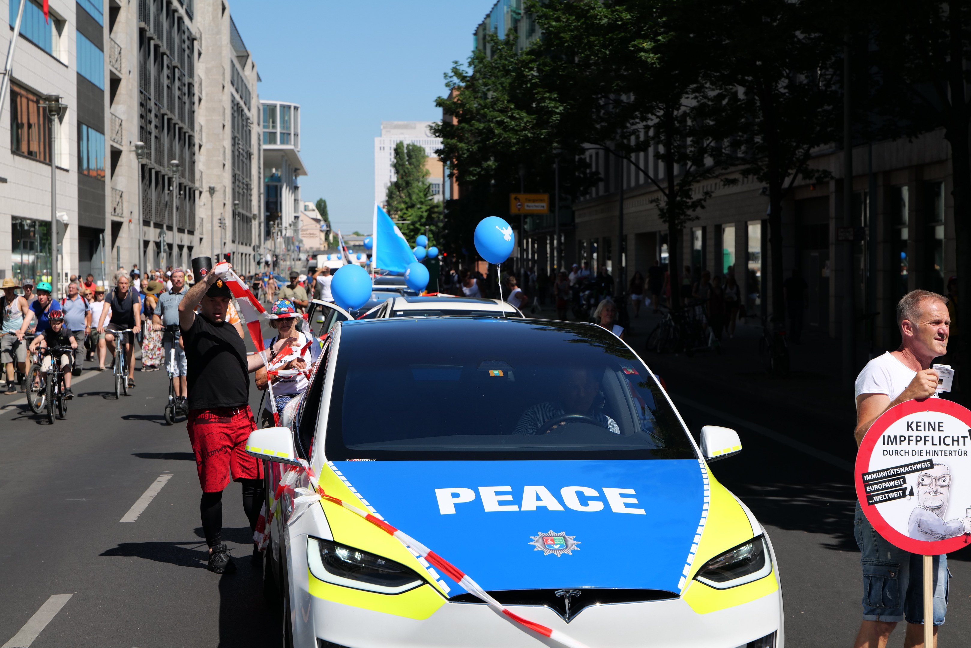Gruppe von Menschen, die auf Fahrrädern eine Straße entlangfahren, mit einem Polizeiwagen im Vordergrund, der Text anzeigt, einer Person, die ein Schild mit einem Bild und Text hält, und Gebäuden, Bäumen und einem klaren Himmel im Hintergrund.