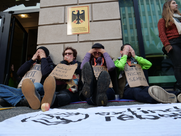 Eine Gruppe von Menschen sitzt vor einem Gebäude auf dem Boden, hält Schilder und hat ein Banner und einen Apfel auf der Straße, sowie eine Frau, die rechts steht und besorgt aussieht.