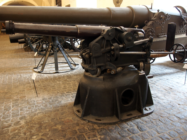 Eine große Gatling-Kanone mit langem Lauf und einem Seitengriff wird in einem Museum ausgestellt, umgeben von Bodenständern, mit einer burgähnlichen Wand mit Fenstern im Hintergrund.