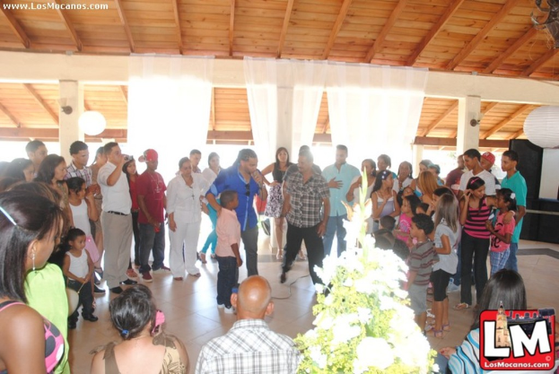 Eine Gruppe von Menschen mit einer Pflanze und einem Logo unten, Vorhänge und ein Lautsprecher im Hintergrund sowie Text in der oberen linken Ecke.