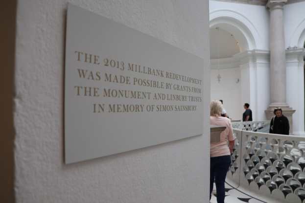 Gruppe von Menschen, die in einem großen, offenen Gebäude mit einer Wandtafel, Säulen, einem Zaun, einer Decke mit Lampen und einem Schild über die 2013er Umgestaltung des Millbank-Geländes stehen, die von den Monument- und Linbury-Stiftungen im Andenken an Simon Sainsbury finanziert wurde.