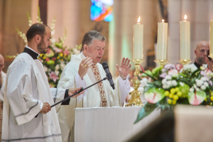 Der Bischof der Kirche des Heiligen Grabes steht an einem Podium mit einem Mikrofon, während er eine Gruppe von Menschen anspricht, mit einem Tisch mit Kerzen und Blumen zu seiner Rechten und Blumensträußen und einem Bleiglasfenster im Hintergrund.