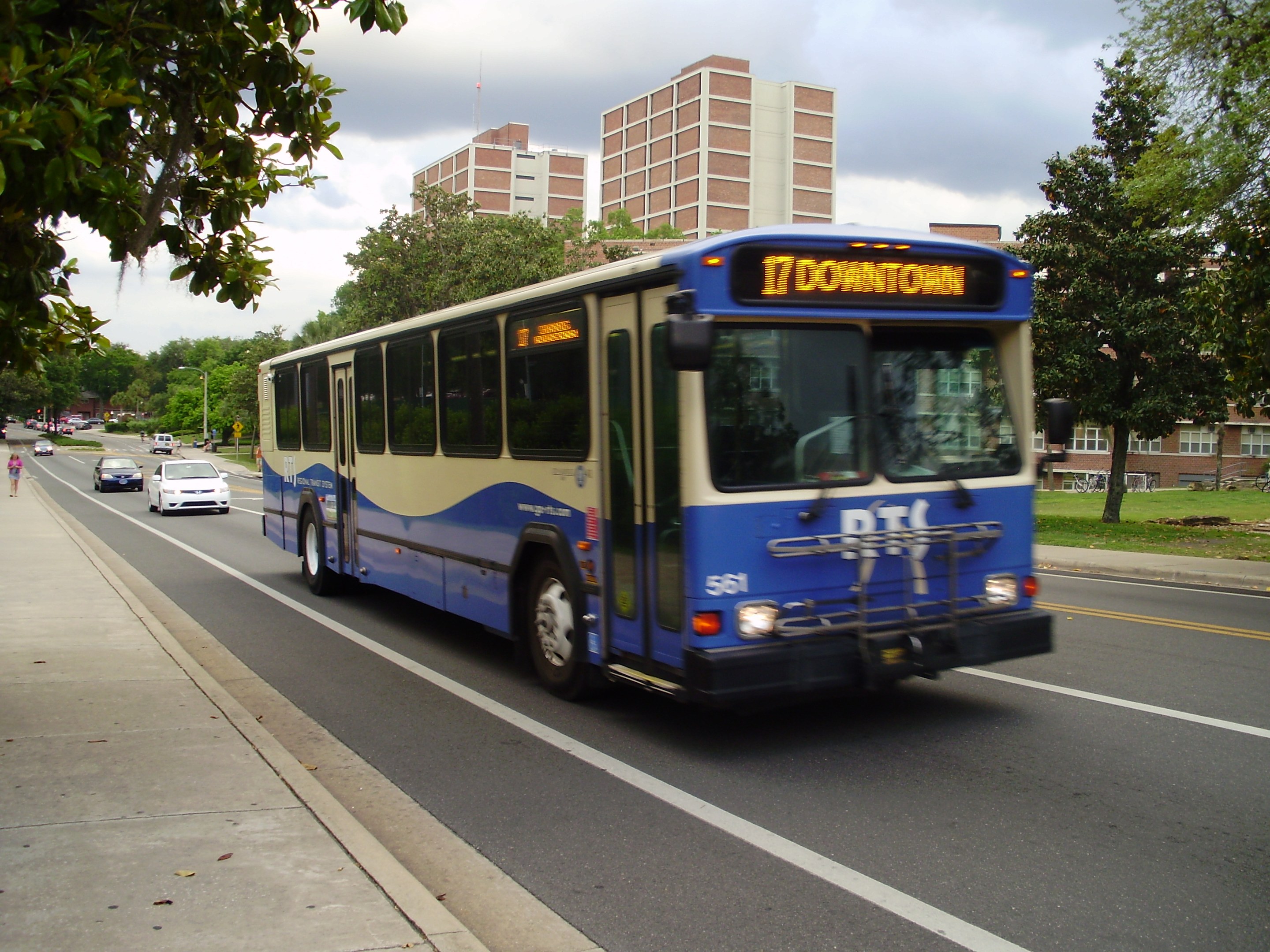 Ein blauer und weißer Shuttlebus fährt eine Straße mit hohen Gebäuden entlang, mit ein paar Fußgängern auf dem Bürgersteig links und Bäumen, Polen und einem klaren blauen Himmel im Hintergrund.
