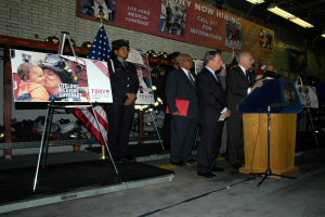 Eine Gruppe von Menschen steht in der Nähe eines Rednerpults mit Mikrofonen, umgeben von Schildern auf Ständern, Flaggen und Schildern an der Wand, alles unter hellen Scheinwerfern.