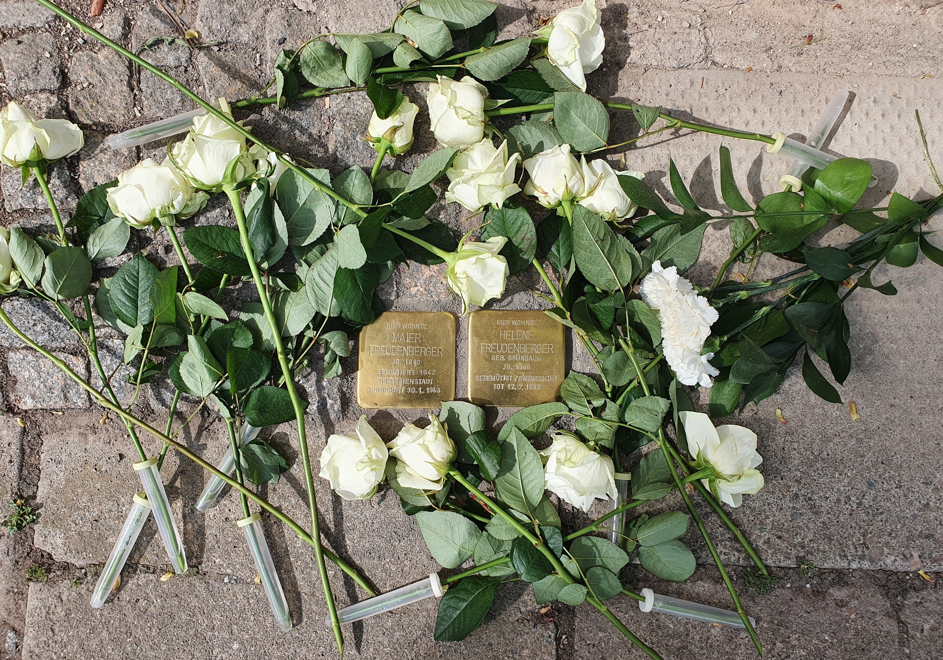 Ein Kranz aus weißen Rosen und zwei goldfarbene Gegenstände mit Inschrift auf dem Boden vor einem Berliner Denkmal.