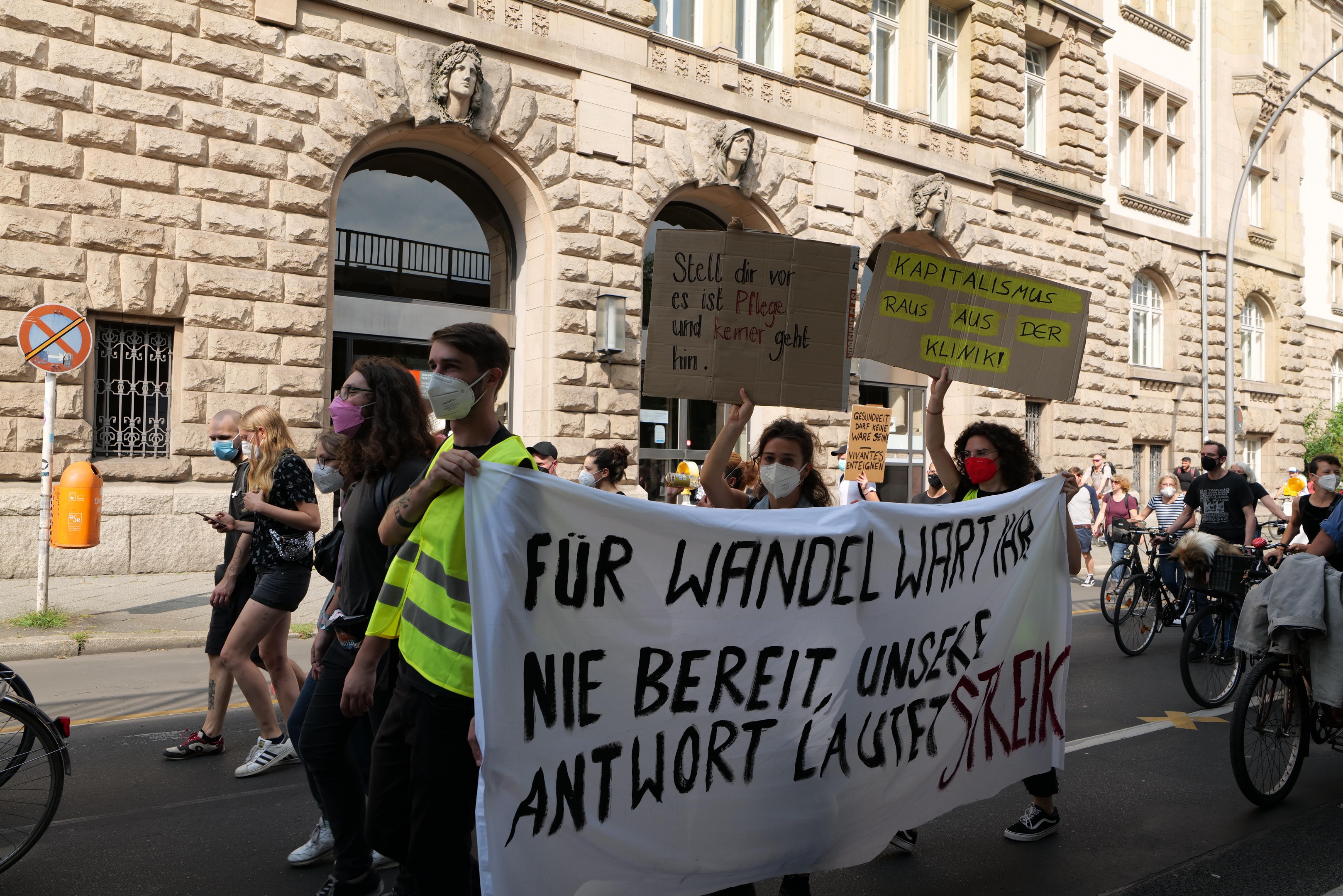 Eine Gruppe von Menschen marschiert durch eine Straße, einige halten Protestschilder und andere fahren Fahrräder, im Hintergrund ein historisches Gebäude mit Rundbogenfenstern, Pfeilern und Skulpturen.