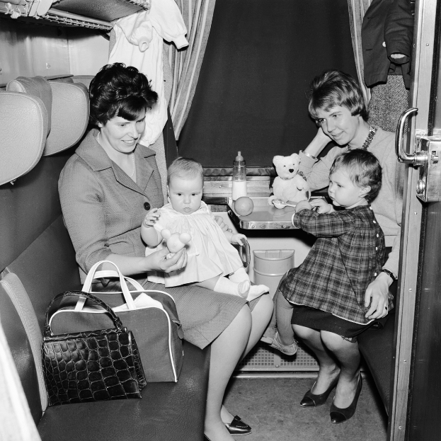 Ein Schwarz-Weiß-Foto von zwei Frauen und zwei Kindern, die in einem Zug sitzen, wobei eine Frau ein Baby hält und die andere zwei Kinder hält, sowie verschiedene Gegenstände auf dem Tisch und dem Sitz.